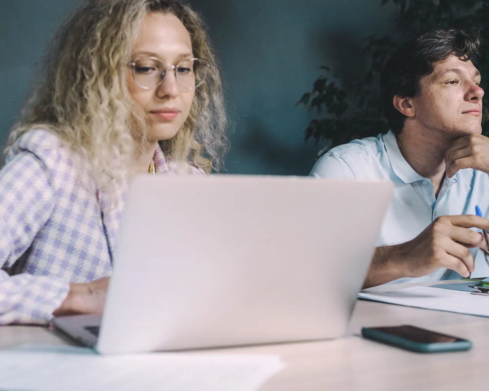 Two colleagues working side by side at a table with a laptop and notes