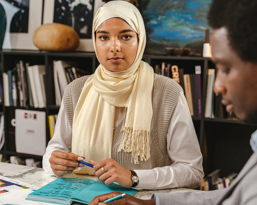 Two colleagues in conversation across a meeting table, one listening closely while the other speaks